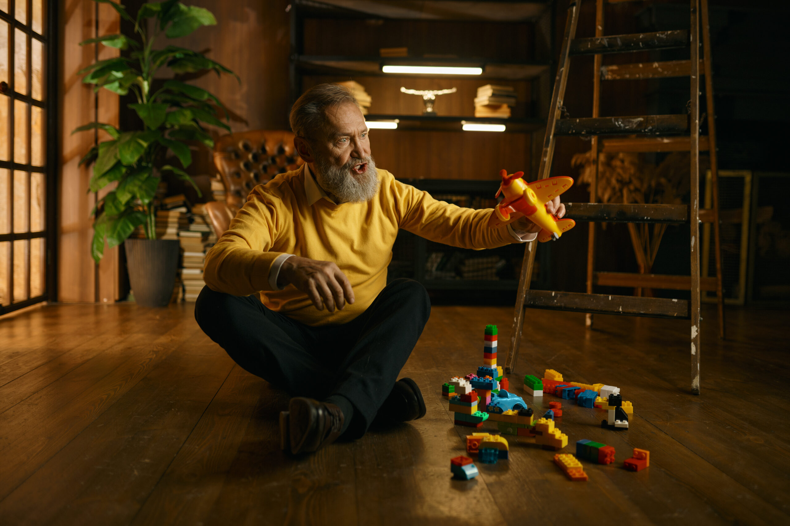 Happy enthusiastic old retired man playing toys while sitting on home floor over cozy eco-friendly interior design